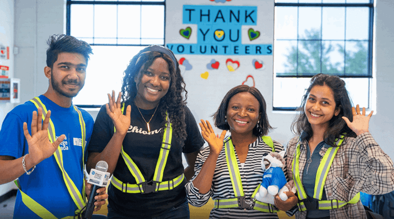 Diverse young students smile and waves at the camera while volunteering